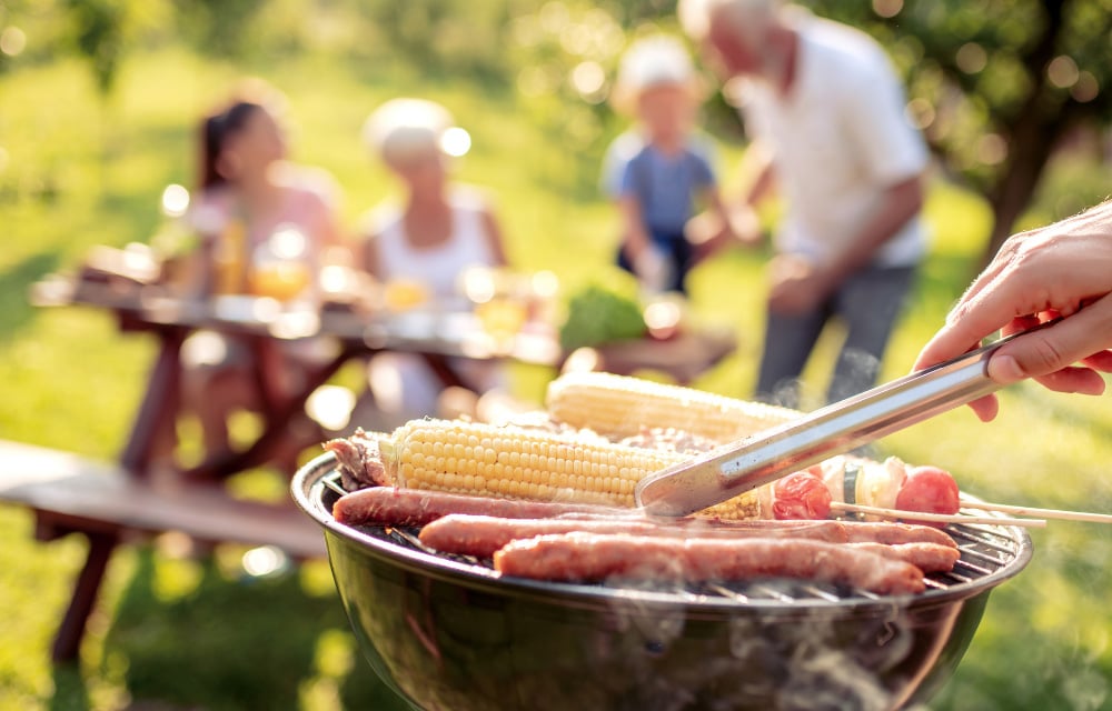 Nahaufnahme eines Grills mit Würstchen und Maiskolben, im Hintergrund eine Familie an einem rustikal dekorierten Gartentisch.