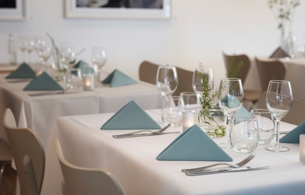 Schlichte, elegante Tischdekoration für eine Betriebsfeier mit weißen Tischdecken, Gläsern und hellblauen Faltservietten.