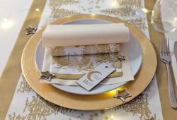 Ein einzelnes, festlich gedecktes Gedeck in Gold und Weiß. Auf dem goldenen Platzteller liegen Servietten, eine davon mit goldenem Waldmotiv. Dazu kleine goldene Sterne als Dekoration.