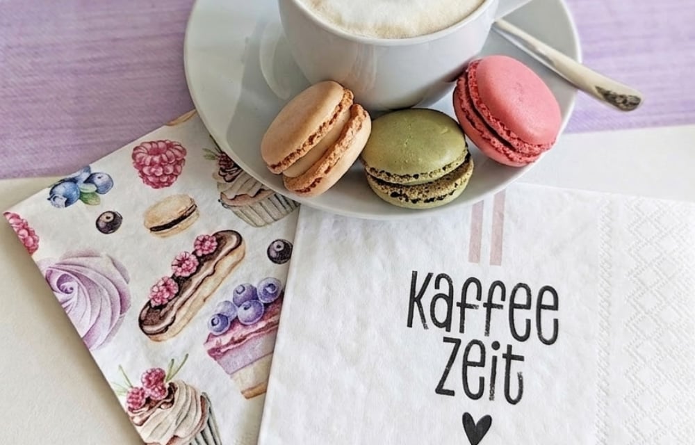Stilvolle Tischdeko zur Kaffeezeit: Bunte Macarons auf einer Untertasse neben dekorativen Servietten mit Gebäck-Motiven.