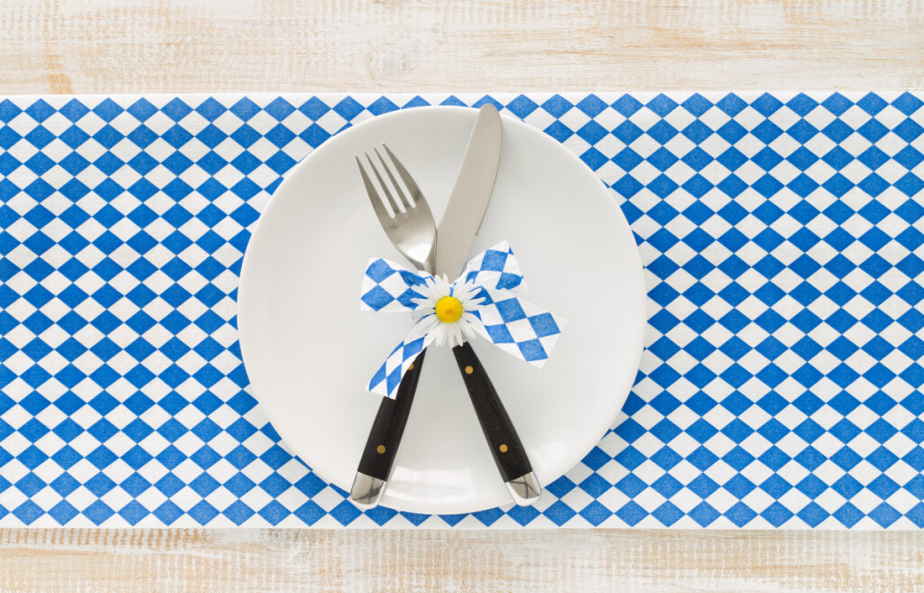 Minimalistische bayerische Tischdeko: Weißer Teller auf Rauten-Tischläufer mit Besteck und einer blau-weißen Schleife.